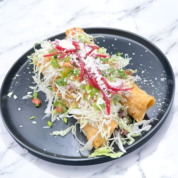 Two Honduran taquitos, topped with shredded cabbage, diced tomatoes, red onions, and crumbled white cheese.