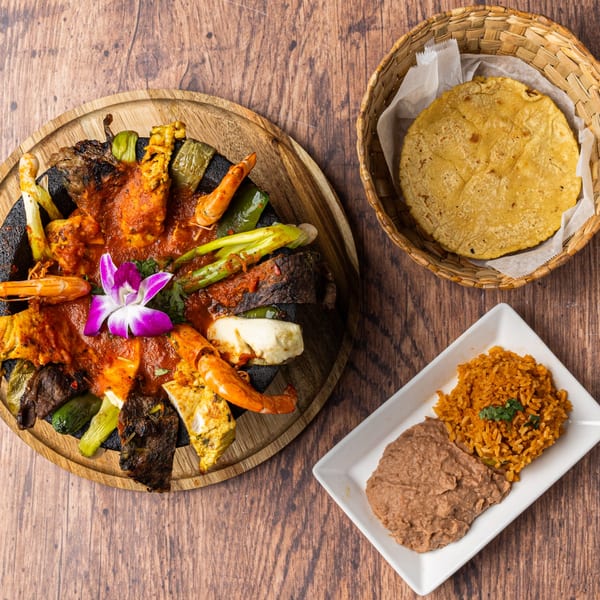 Honduran molcajete with grilled meats, shrimp, vegetables, and a purple orchid, served with rice, beans, and tortillas.