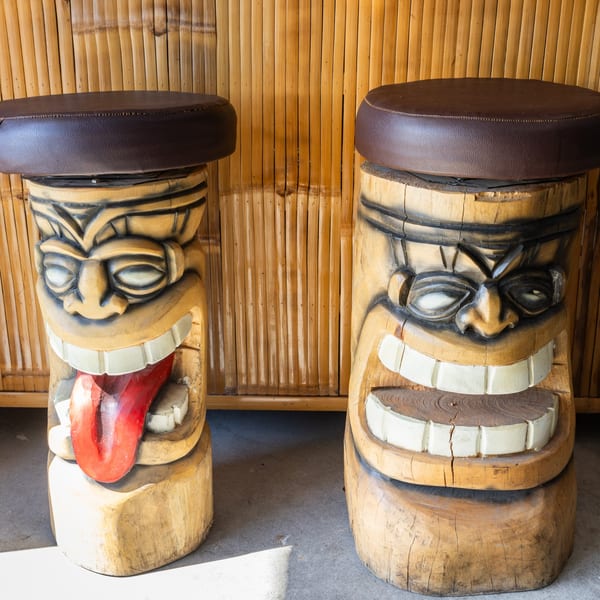 Two wooden tiki bar stools with dark brown cushions against a bamboo wall.