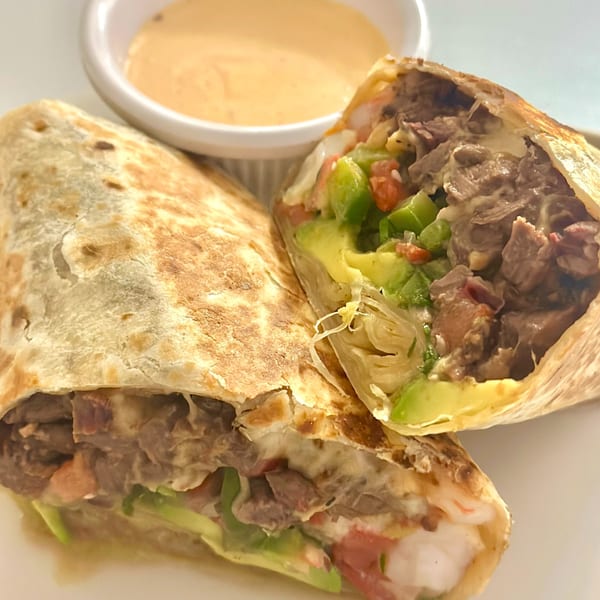 Two halves of a Honduran burrito filled with meat, avocado, and vegetables, with a side of sauce.
