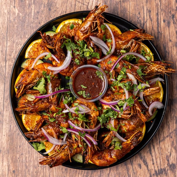 A black plate of Honduran-style grilled shrimp with lemon, red onion, cilantro, and a dipping sauce.