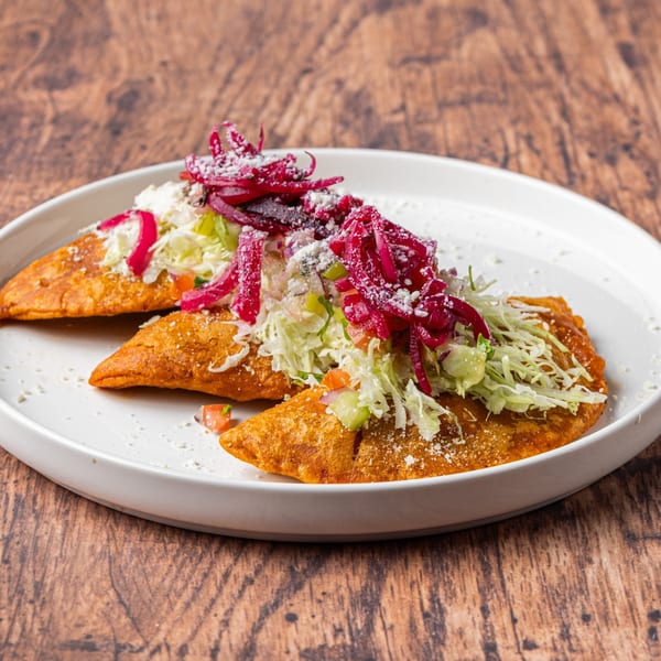 Three golden-brown Honduran pastelitos topped with shredded lettuce, pickled red onions, and crumbled cheese.