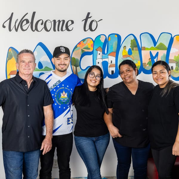 Five smiling people, three women and two men, standing in front of a colorful mural that says "Welcome to La Champa".