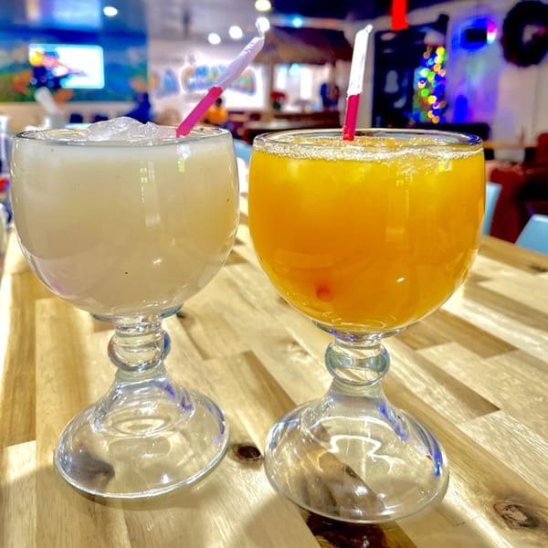Two La Champa Latin Grill drinks, one light and one orange, with straws on a wooden table.