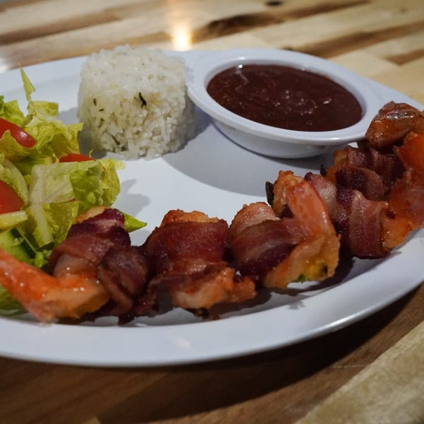 Bacon-wrapped shrimp with rice, salad, and a side of dark sauce on a white plate.